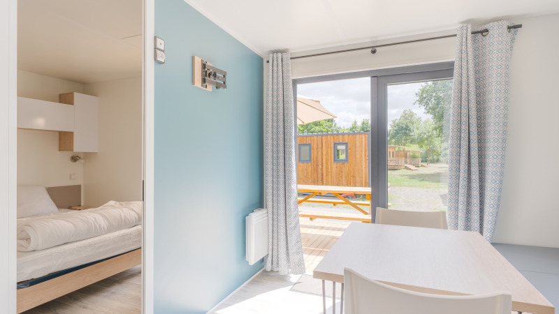 Interior luminoso de lodge con dormitorio, mesa de comedor, ventanales con cortinas y vista a la terraza.