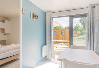 Intérieur lumineux de lodge avec chambre, table à manger, grandes fenêtres, rideaux et vue sur terrasse.