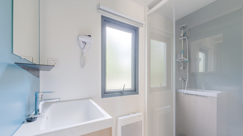 Bright, modern bathroom at a glamping site featuring a shower, window, sink, and wall-mounted hairdryer.