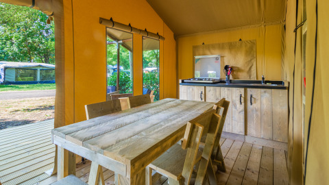 Binnenzicht van een safaritent met houten tafel, stoelen, een kitchenette en zicht op het groen buiten.