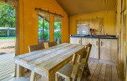 Binnenzicht van een safaritent met houten tafel, stoelen, een kitchenette en zicht op het groen buiten.