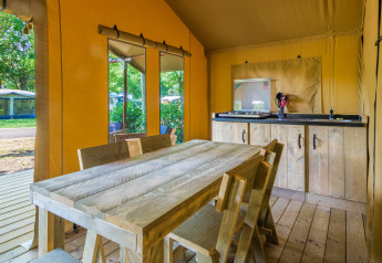 Interior of a safari tent featuring a wooden dining table, chairs, kitchenette, and views of greenery outside.