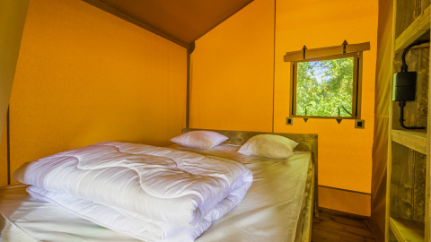 Interior de una tienda safari con cama doble, sábanas blancas y ventana con vistas al exterior natural.