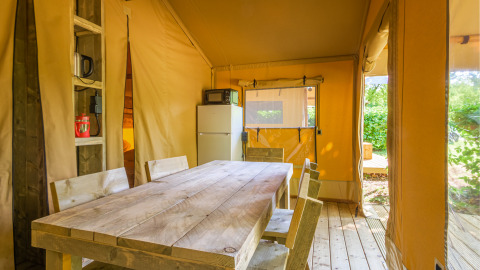 Binnenkant van een safaritent met houten tafel, stoelen, koelkast en ramen met zicht op de natuur.