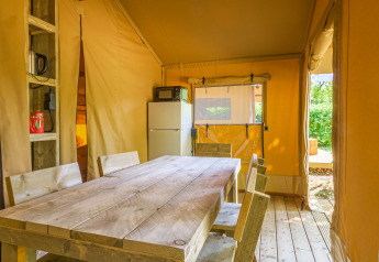 Interno di una tenda safari con tavolo e sedie in legno, frigorifero e finestre che danno sulla natura.