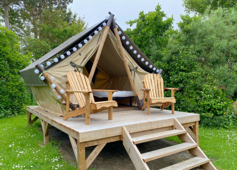 Glamping tent on a raised wooden platform with two wooden chairs, set amid lush green foliage outdoors.