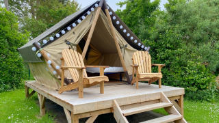 Glamping tent on a raised wooden platform with two wooden chairs, set amid lush green foliage outdoors.