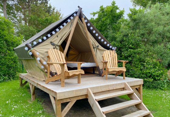 Glamping tent on a raised wooden platform with two wooden chairs, set amid lush green foliage outdoors.