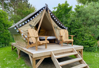 Glamping tent on a raised wooden platform with two wooden chairs, set amid lush green foliage outdoors.