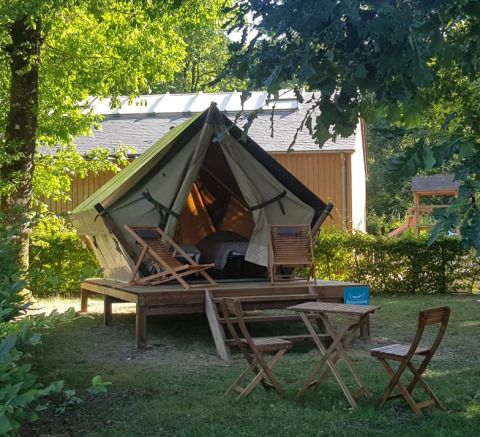 Tenda safari su piattaforma in legno con sdraio e tavolo, immersa nel verde vicino a una casetta.