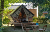 Tenda safari su piattaforma in legno con sdraio e tavolo, immersa nel verde vicino a una casetta.