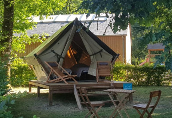 Tenda safari su piattaforma in legno con sdraio e tavolo, immersa nel verde vicino a una casetta.