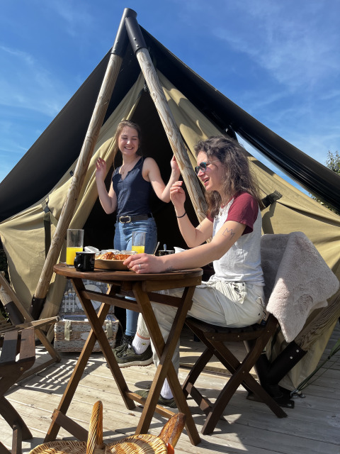 Dos mujeres jóvenes disfrutan del desayuno y el sol frente a una tienda glamping con mobiliario de madera.
