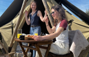 Dos mujeres jóvenes disfrutan del desayuno y el sol frente a una tienda glamping con mobiliario de madera.
