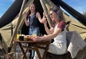 Dos mujeres jóvenes disfrutan del desayuno y el sol frente a una tienda glamping con mobiliario de madera.