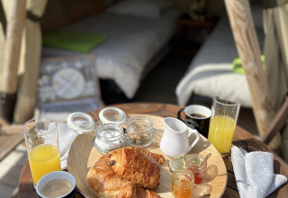 Desayuno con croissants, jugo y café en una mesa pequeña frente a una tienda glamping con dos camas.