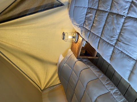 Deux lits simples avec couettes grises et une lanterne sur une table de nuit dans une tente de glamping.