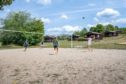 Four people playing beach volleyball on a sandy court at a holiday park with cabins and green landscape.