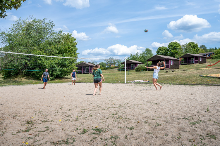 Vier Personen spielen Beachvolleyball auf einem Sandplatz, Ferienhütten und Natur im Hintergrund.