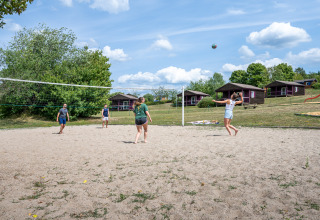 Fire personer spiller beachvolleyball på en sandbane ved feriepark, med hytter og grønt landskab bagved.