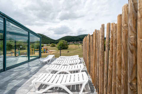 Terraza exterior con tumbonas junto a una pared de cristal y valla de madera en Camping Seasonova Les Vosges du Nord.