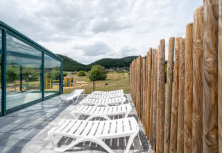 Terrasse mit weißen Liegestühlen, Glaswand und Holzzaun im Camping Seasonova Les Vosges du Nord, Frankreich.