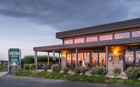 Evening view of Huttopia Les Falaises - Normandie holiday park in France, with guests at a warmly lit café.