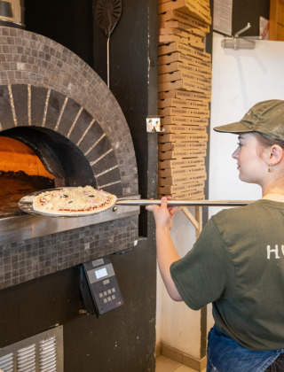 Un empleado horneando pizza en horno de piedra en Huttopia Les Falaises - Normandie, parque vacacional.