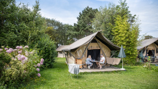 Twee mensen zitten voor een safaritent genaamd Tente Canadienne, omgeven door groen en bloemen op de camping.