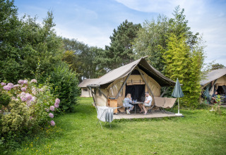 To personer sidder foran en safari Tente Canadienne omgivet af grønt og blomster på en campingplads.