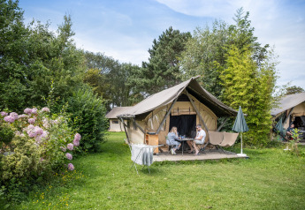 To personer sidder foran en safari Tente Canadienne omgivet af grønt og blomster på en campingplads.