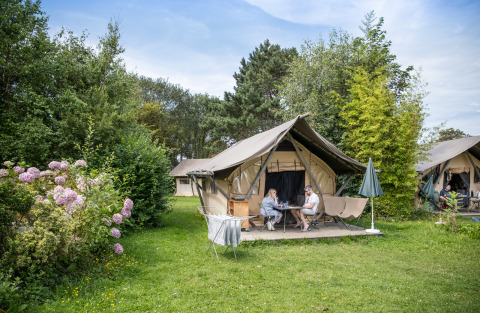 Two people sit outside a safari tent named Tente Canadienne, surrounded by greenery and flowers at a campsite.