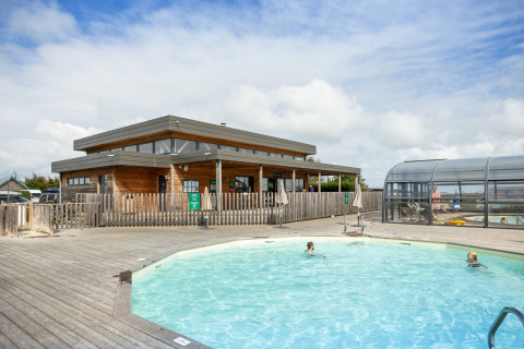 Piscina exterior con bañistas y edificio de madera moderno en Huttopia Les Falaises, Normandía, Francia.