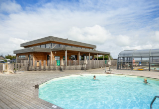 Buitenzwembad met bezoekers en een modern houten gebouw bij Huttopia Les Falaises in Normandië, Frankrijk.