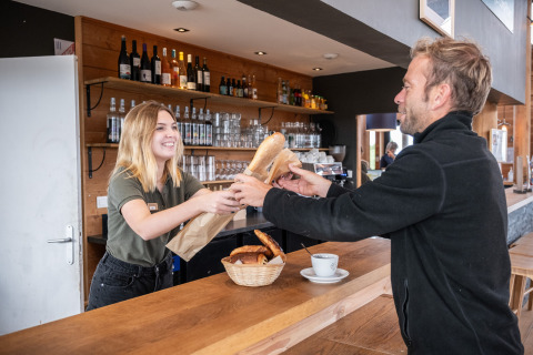 Cliente recibe una baguette fresca de una empleada sonriente en Huttopia Les Falaises - Normandía, Francia.