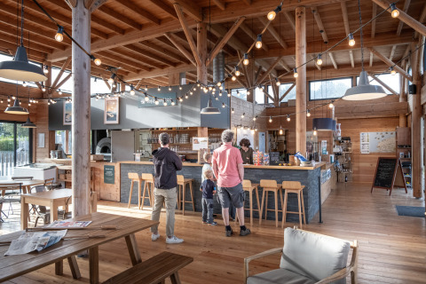 Bezoekers bestellen aan de toog van het café in het houten interieur van Huttopia Les Falaises, Normandië.
