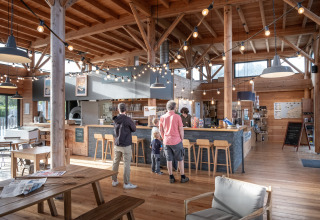 Bezoekers bestellen aan de toog van het café in het houten interieur van Huttopia Les Falaises, Normandië.