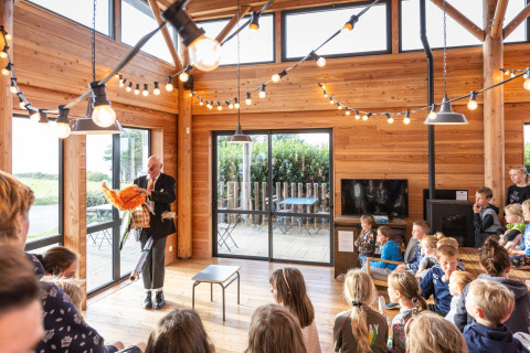 Een man entertaint kinderen in een houten zaal met sfeerverlichting bij Huttopia Les Falaises, Normandië.