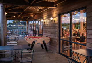 Abendaufnahme einer Holzterrasse mit Tischfußball und Sitzplätzen im Ferienpark Huttopia Les Falaises, Normandie.