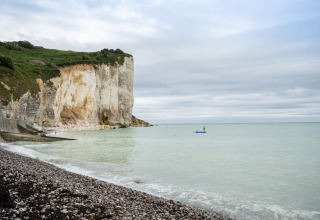 Coastal cliffs by Huttopia Les Falaises in Normandy, France, with pebble beach and calm sea under clouds.