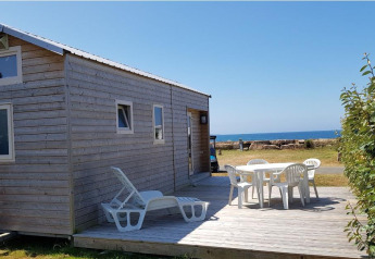 En lille træhus kaldet Tiny House med terrasse, havudsigt og havemøbler under en klar blå himmel.