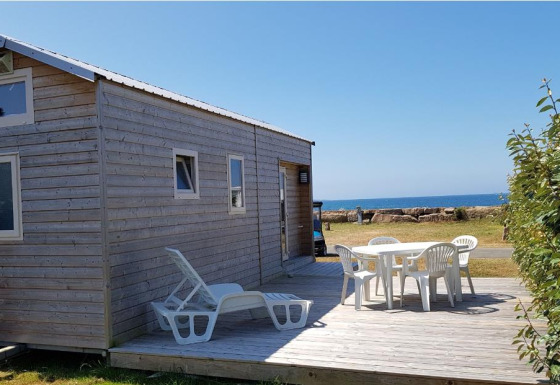 Casa pequeña de madera llamada Tiny House con terraza, sillas blancas y vista al mar bajo cielo azul.
