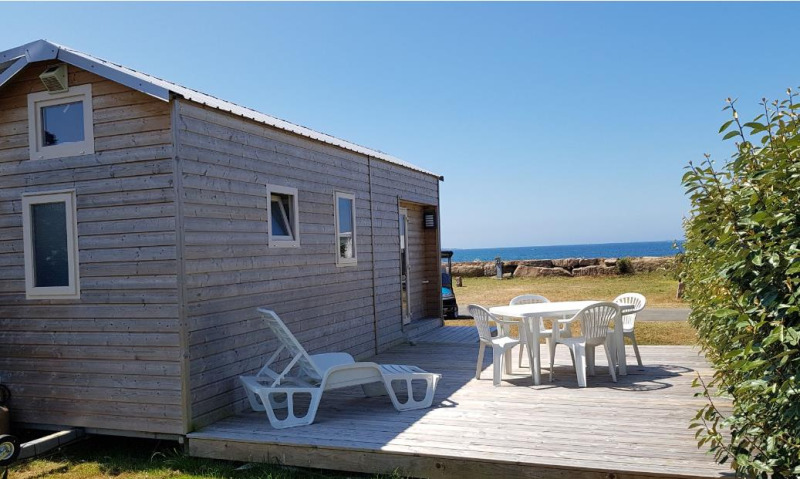 Tiny House wooden cabin with outdoor terrace, plastic chairs, and sea view under a clear blue sky.