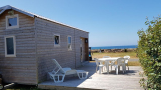 Casa pequeña de madera llamada Tiny House con terraza, sillas blancas y vista al mar bajo cielo azul.