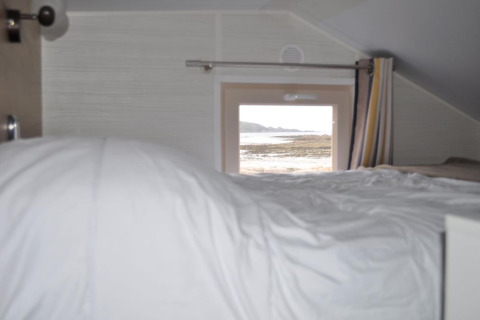 Vista interior de una tiny house con cama y ventana hacia la playa en Camping Seasonova Les 7 Iles, Francia.