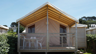 Cabina Eco Lodge con struttura in legno, veranda coperta e sedie bianche, circondata dal verde.