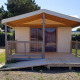Ecolodge Pmr kabine med rampe og træterrasse, omgivet af grønne hække under en blå himmel.