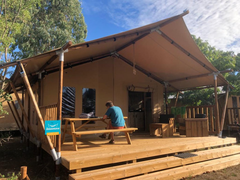A person sits at a picnic table on a wooden deck outside a safari tent, surrounded by trees and sky.