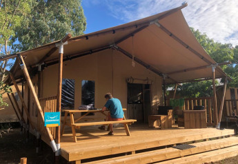 A person sits at a picnic table on a wooden deck outside a safari tent, surrounded by trees and sky.