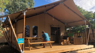A person sits at a picnic table on a wooden deck outside a safari tent, surrounded by trees and sky.